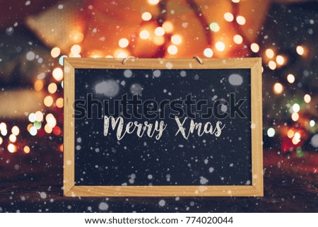 merry xmas written in english language on a chalkboard. trendy xmas greeting card, ideal for holiday season. bokeh lights in the background. snowflake effect. snow falling.
