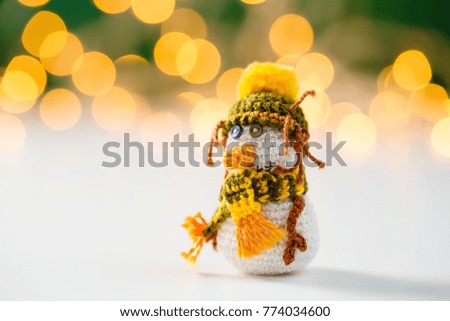 Handmade crochet snowman toy on white background with garland lights bokeh. Christmas still life. Greeting card concept with copy space