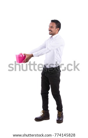 Full-length image of a cheerful guy looking to the side is wearing white shirt, black trousers, and boots giving Christmas gift. Isolated on white background.
