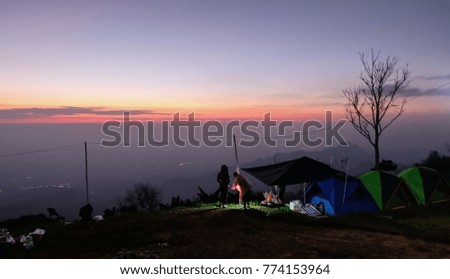 Silhouette of  peoples standing  and watching sunrise beside camp and tents in the morning. On the background misty sky, mountains and tree.