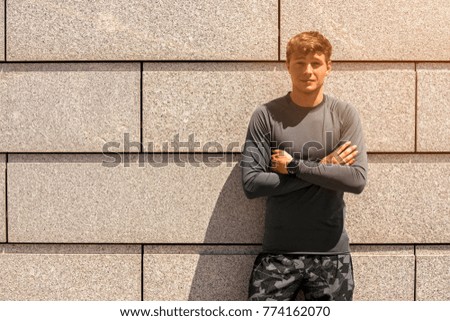 Muscular build young runner crossed arms, tired after training outdoors, athletic jogger in bright sportswear resting after run on beautiful concrete wall background, fitness concept
