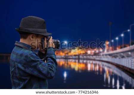 A cute boy wearing a black hat is taking a picture of the city waterfront lights in the night.