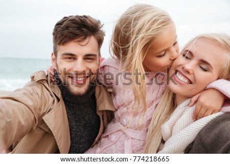 Portrait of a joyful family with a little daughter taking a selfie together while standing at the beach