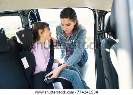 Woman helping her daughter to fasten seatbelts in the car, the girl is sitting on a safety child car seat Royalty-Free Stock Photo #774241534