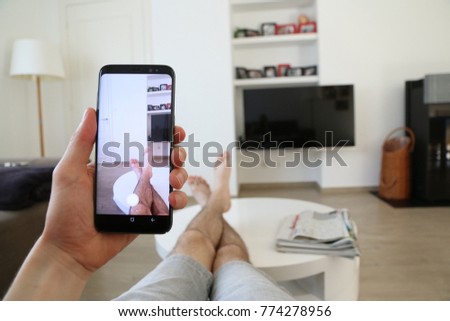 A hand holding a modern generic mobile phone which displays the camera app on the touch screen. The person's legs are next to newspapers, on the table, in front of the TV. A concept of taking pictures