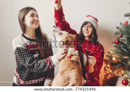 Happy holidays concept, family New Year celebration, Woman opening Christmas present near tree with sister, cute smiling girls in stylish warm sweaters playing with dog near Christmas tree