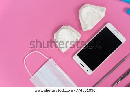 on the pink surface lie a white smartphone, a medical face mask, a plaster cast of teeth and dental tools