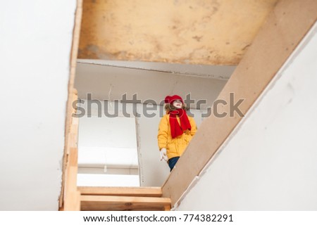 A rectangular opening of the second floor of the house under construction, in which a girl is seen in a winter yellow jacket, a red knitted hat and a scarf