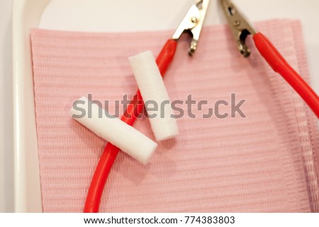 Two dental cotton rolls resting on a pink dental bib and red rubber neck strap with silver alligator clips resting on a white medical tool tray. 