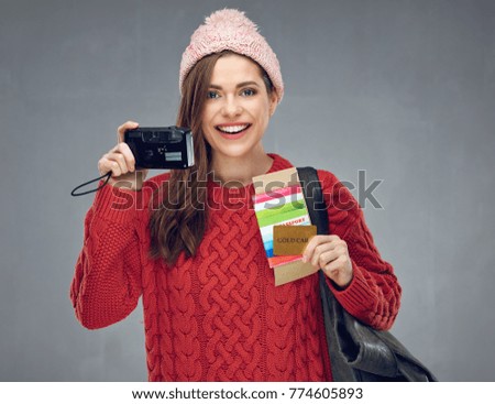 Happy woman dressed red knitted sweater holding camera, credit card with passport and tickets. Gray wall back.