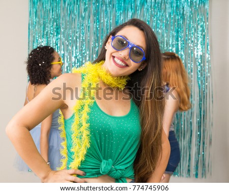 Oriental woman is wearing blue sunglasses and yellow scarf. In the background friends are dancing. Carnival in Brazil