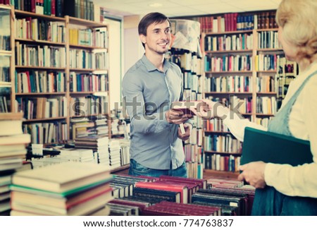 Young glad smiling  man taking chosen book from seller in book shop