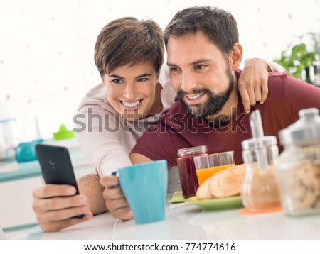 Young cheerful couple having breakfast at home together and taking selfies using a smartphone