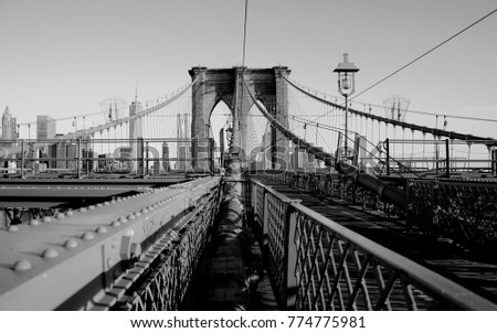 A black and white photograph of the Brooklyn Bridge from the perspective of suspension lines. The Manhattan skyline is in the background.