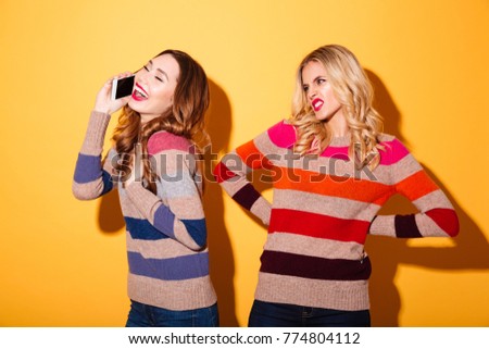 Portrait of a happy girl talking on mobile phone while her angry girlfriend looking at her isolated over yellow background