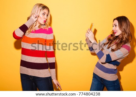 Cheery girl taking a photo of her girlfriend isolated over yellow background