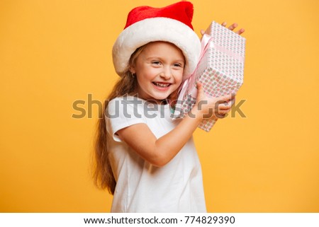 Picture of little laughing girl standing isolated over yellow background wearing christmas santa hat. Looking aside holding surprise gift box.