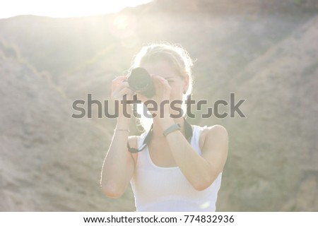 A blonde woman taking pictures on a hiking trip. 
White tank top, black camera. 
Bright sun coming up from behind the hills. 