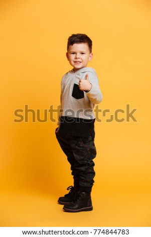 Picture of cheerful cute little boy child standing isolated over yellow background. Looking camera showing thumbs up.