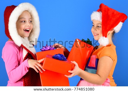 Children with happy faces give presents on blue background. Girls celebrate New Year, copy space. Childhood, leisure and xmas sale concept. Kids in Christmas hats with big gift boxes open presents.