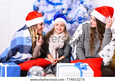Girls with happy faces with white and blue Christmas tree on background. Sisters hang out by gift boxes. Christmas morning and New Year concept. Family or friends in Santa hats near present boxes