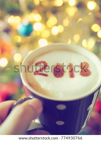 Hand holding a cup of coffee with the number 2018 on frothy surface, bokeh light background. Holiday food concept image for New Year 2018. (selective focus, vintage filter, space for text)
