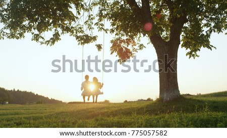 LENS FLARE SILHOUETTE: Young couple in love swaying on tree swing at golden summer sunset. Happy man and woman swinging under tree in sunny evening. Boyfriend and girlfriend hugging on romantic date. Royalty-Free Stock Photo #775057582