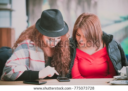 Two cute beautiful young women friends sitting on coffee shop bench on summer day, browsing pictures on smartphone, back view, focus on device screen