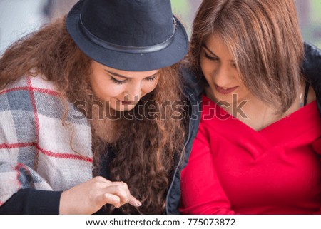 Two cute beautiful young women friends sitting on coffee shop bench on summer day, browsing pictures on smartphone, back view, focus on device screen