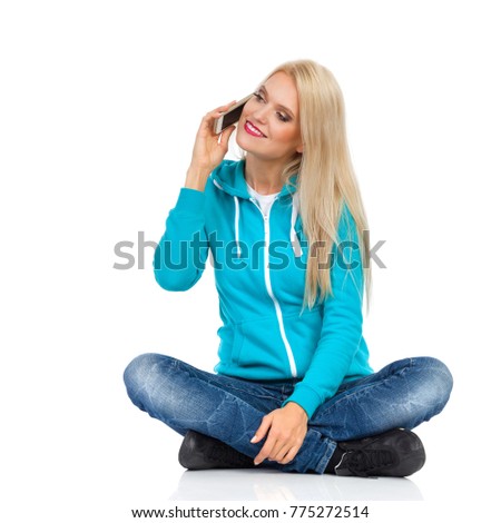 Beautiful blond woman in turquoise sweater and jeans is sitting legs crossed on floor, holding cell phone, looking away and smiling. Front view. Full length studio shot isolated on white.