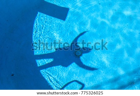 The shadow silhouettes of a diving board and a person with raised arms standing at the edge stand out on the blue, rippled surface of a swimming pool. Royalty-Free Stock Photo #775326025
