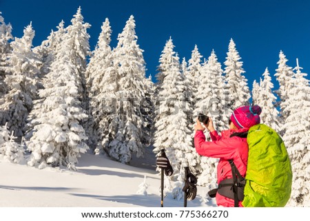 Caucasian young woman in colorful outfit taking a break from backcountry skiing to photograph snow covered Christmas trees