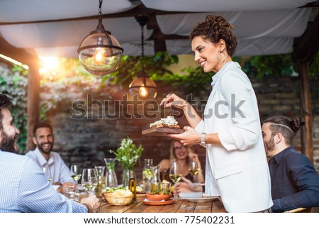 Beautiful Caucasian smiling woman putting a plate with meal on dining table at backyard celebration.
