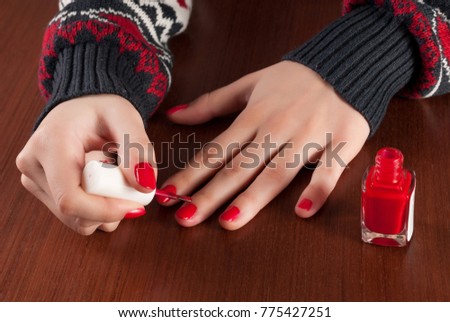 Young girl in sweater paint nails in red color and bottle on wooden desk. Close up