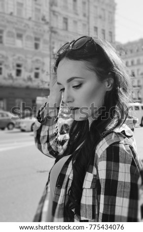 Street portrait of a beautiful brunette woman in a plaid shirt. Sunglasses on the head. Reflection of the sky in glasses glasses. Inviting look of a young woman. Mysterious smile.