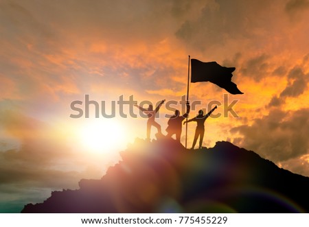 Conquest of height, silhouettes of three people, on top of a mountain, with a flag. Conceptual design. Against the background of the evening sky at sunset with clouds. Royalty-Free Stock Photo #775455229