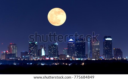 Night view of the Tampa Florida skyline showing skyscrapers with lights and a huge full moon in the sky over the buildings.