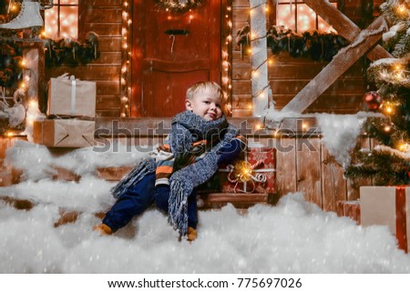 Cute little boy in winter clothes sits on the porch of the magic house of Santa Claus with gift box. Miracle time. Merry Christmas and Happy New Year.