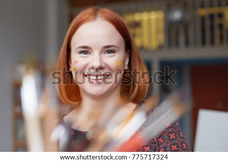 Close up portrait of redheaded female painter captured in time of creative process in art workplace. Head shot of ginger artist with blur brushes on first plan of picture. Positive face expressions.