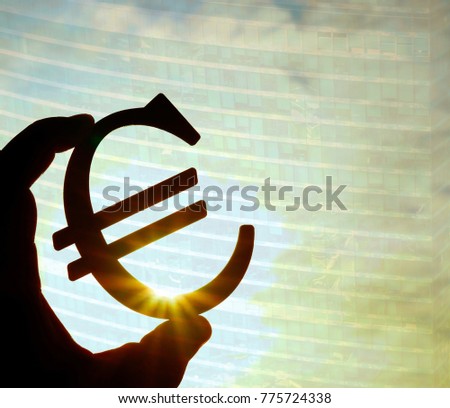 man's hand hold the Euro icon silhouette against sunny blue, skyscraper  and office building. sun rays. euro sign, symbol of money, idea of Euro Union