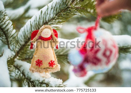 male hand hanging decorative round knitted toy ball near christmas angel on snowy Christmas tree branch background. 