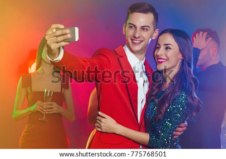 Photo from the festive party. Beautiful young couple with a background of dancing friends taking pictures of themselves on the phone or making selfies.
