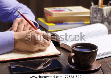 Male hand with red pencil writes in notepad, opened book, smartphone, black coffee in brown cup on dark wood working table, working or study concept.