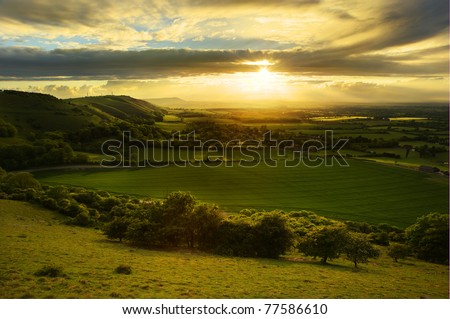Lovely landscape of countryside hills and valleys with setting sun lighting up side of hills whit sun beams through dramatic clouds Royalty-Free Stock Photo #77586610