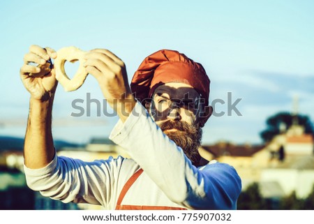 Handsome bearded man cook chef in uniform and red hat and apron with long beard standing with dough in shape of heart sunny day outdoor on blue sky background