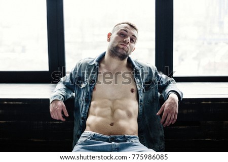 Confident young man in unbuttoned denim jacket posing white sitting near window