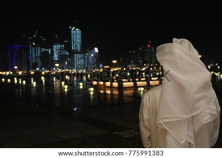 An Arab Qatari man wearing a white thobe or thwab (Arab garment) and white ghutra (or keffiyeh or headress) looking out to the evening or night skyline of Doha, Qatar Royalty-Free Stock Photo #775991383