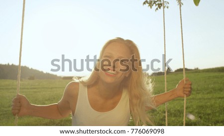 LENS FLARE, PORTRAIT: Young woman glowing in spring afternoon sunshine while happily swaying on fun tree swing. Smiling girl glows in summer sunrays and radiates with joy as she swings in vast meadow.