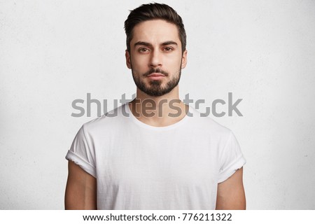 Unshaven confident serious young male enterpreneur with attractive appearance dressed casually as spends weekends at home, isolated over white concrete background. Stylish handsome guy indoor Royalty-Free Stock Photo #776211322