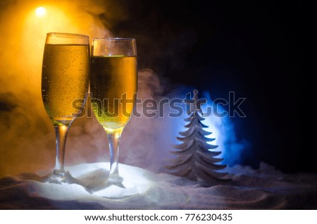 New Year Eve celebration background with pair of flutes and bottle of champagne with Christmas attributes (or elements) on snowy dark toned foggy background. Selective focus. Useful as greeting card